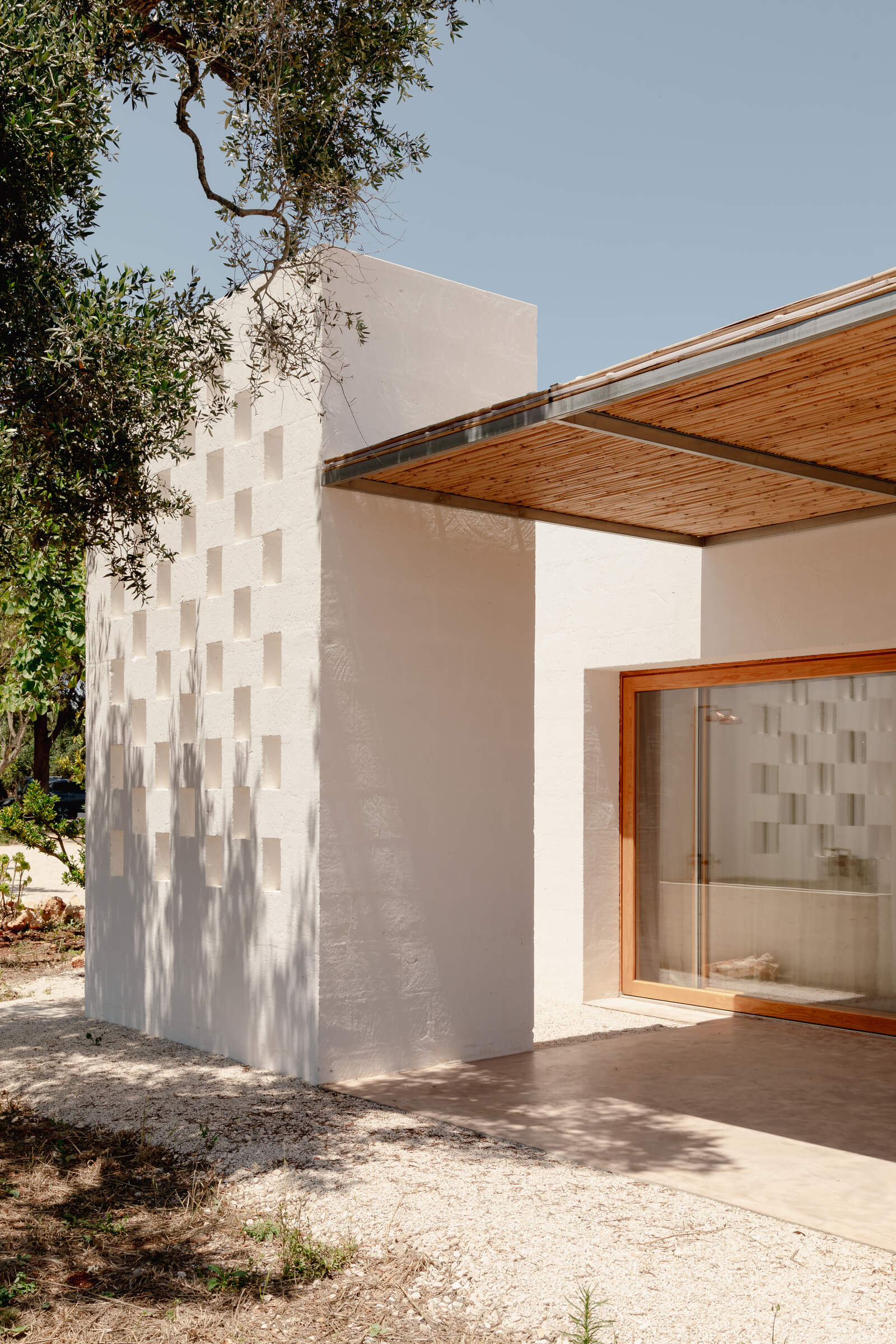 Casanguilla, Europe — Minimalist white-rendered villa with geometric window pattern, wooden pergola, and olive tree framing modernist courtyard architecture in Mediterranean sunlight.