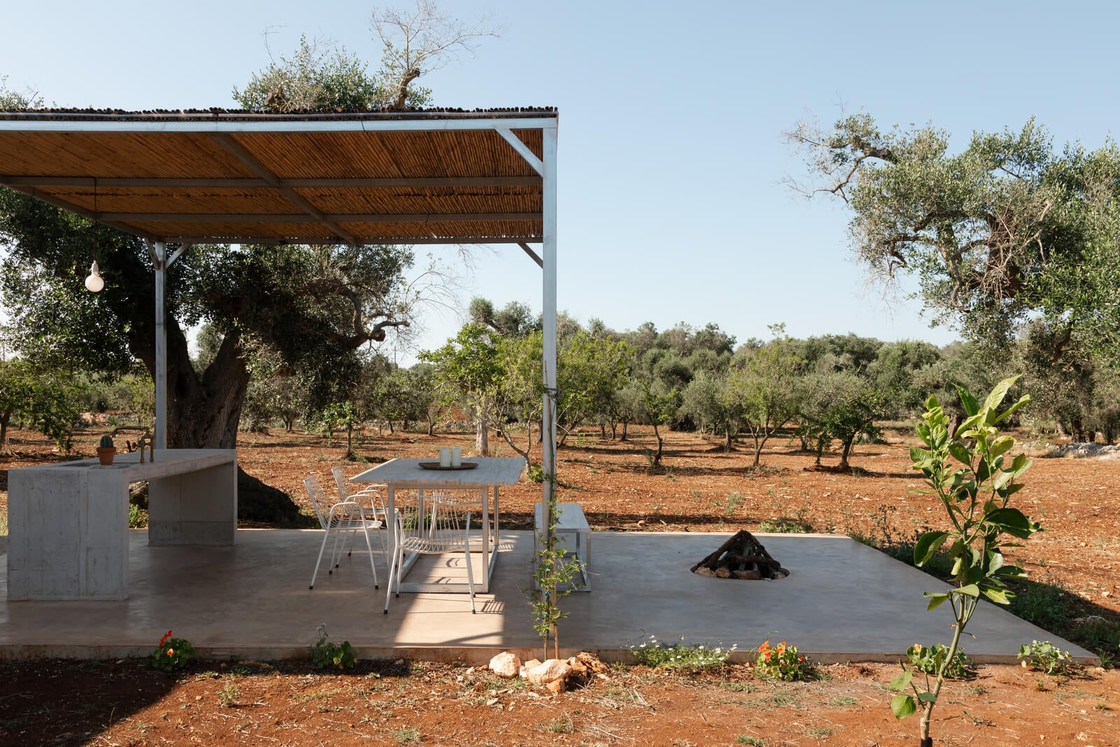 Casanguilla, Europe — Concrete terrace with wooden pergola overlooking olive grove, minimalist outdoor dining setup with fire pit, Mediterranean rural landscape.