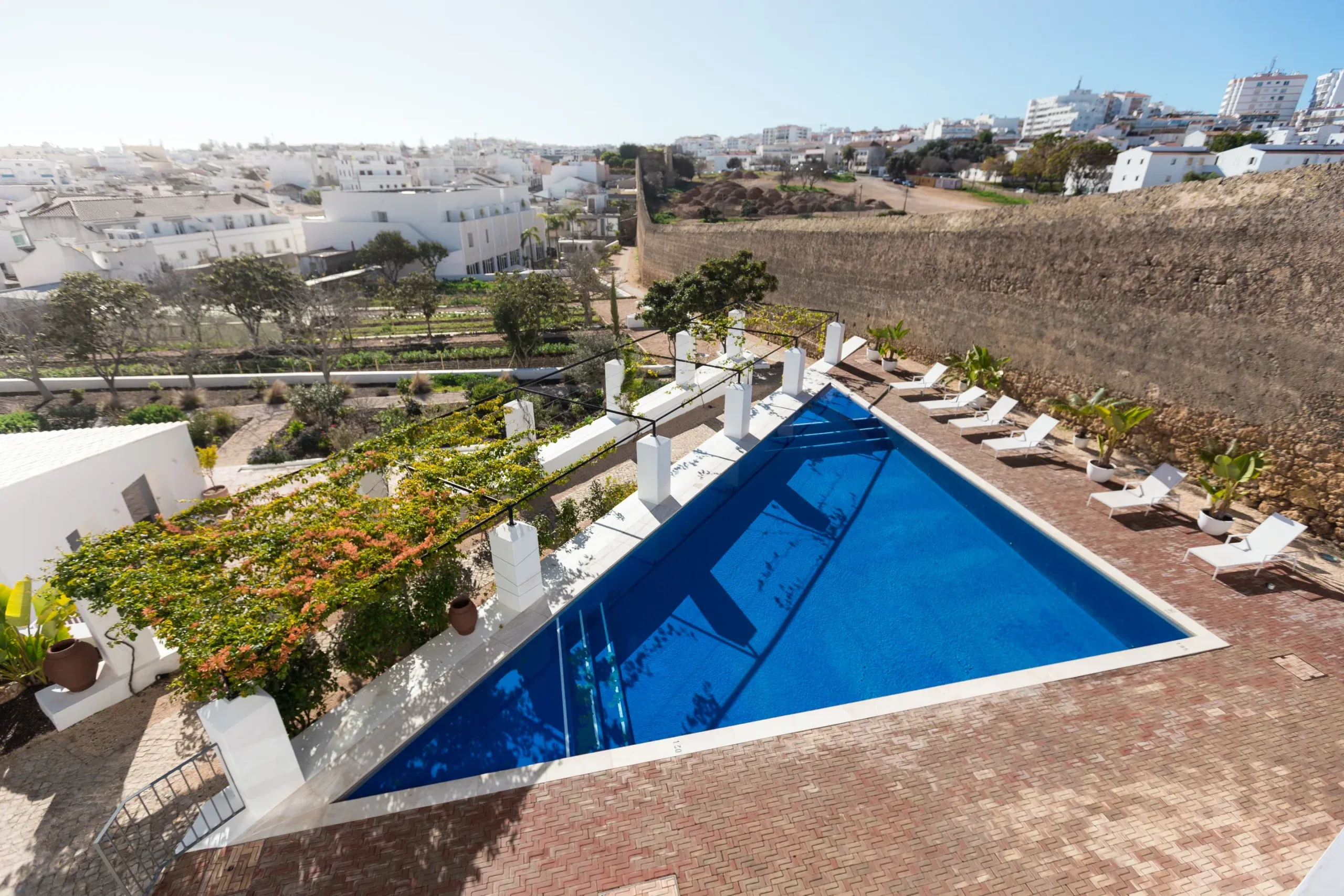 Casa Mãe, Europe — Angular blue swimming pool with white loungers, terraced gardens, and whitewashed architecture overlooking hillside town under clear sky.