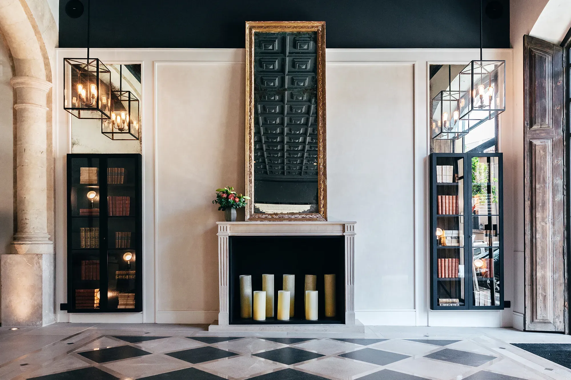 Sant Francesc Hotel Singular, Balearic Islands, Spain — Neoclassical fireplace with black marble surround, gilt-framed mirror, flanking display cabinets, geometric floor, brass pendants.