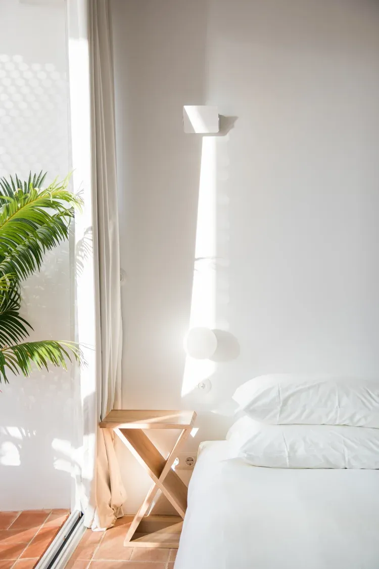 Casa Mãe, Europe — Minimalist bedroom with natural light streaming through tall window, white bedding, wooden side table, potted palm, terracotta flooring, and clean architectural lines.