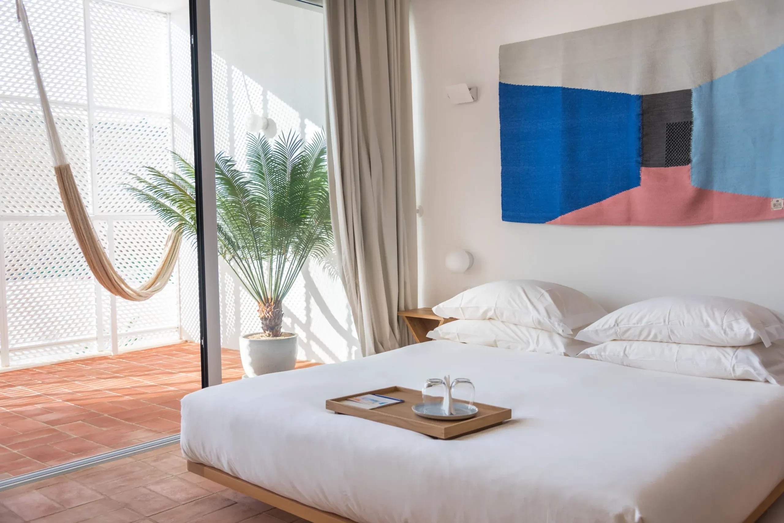 Casa Mãe, Europe — Minimalist bedroom with white bedding, geometric blue and coral artwork, terracotta tiles, and ocean-view balcony with palm plant and hammock.
