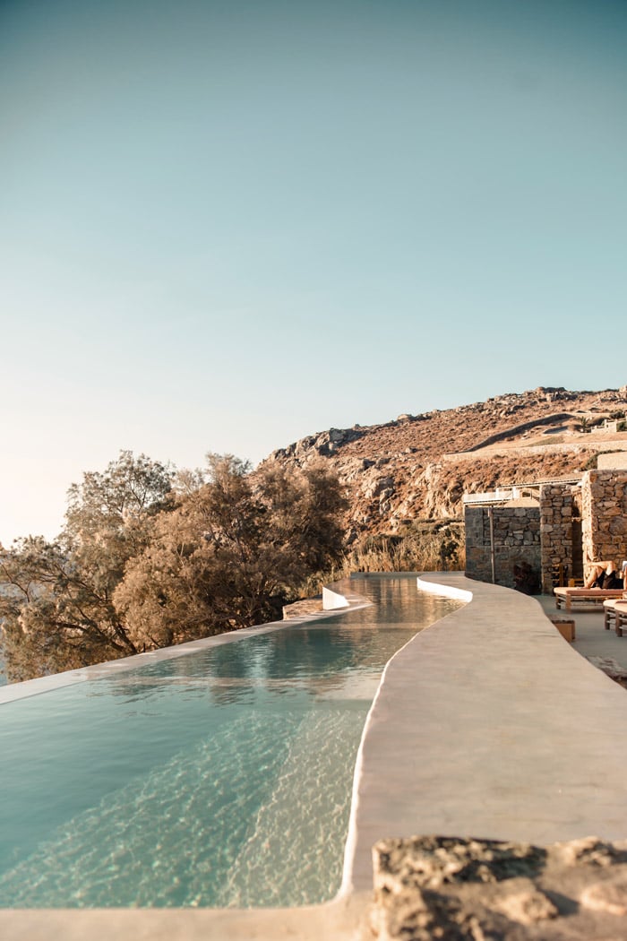 The Wild, Europe — Infinity pool with turquoise water curves along stone terrace overlooking rocky hillside at golden hour, minimalist architecture and desert landscape.