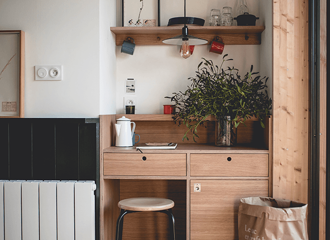 Le Barn, Europe — Natural wood cabinetry with open shelving, potted plant, and minimal black accents beside white radiator in bright Nordic-inspired interior.