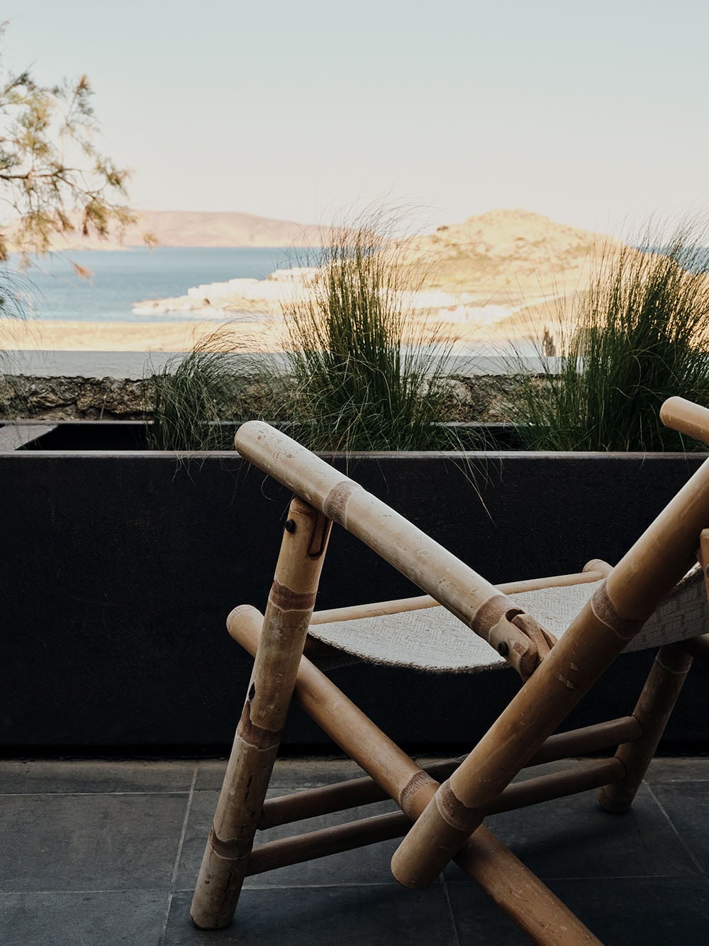 The Wild, Europe — Bamboo lounge chair with canvas seat overlooking coastal dunes and turquoise waters at sunset.