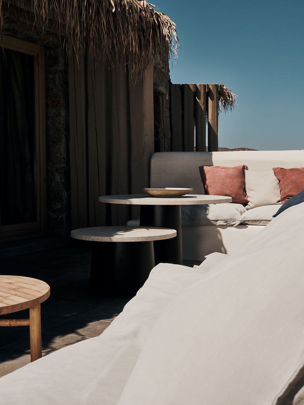 A rooftop lounge at The Wild with pale upholstered seating, dark side tables, thatched structures, and clear Mediterranean light.