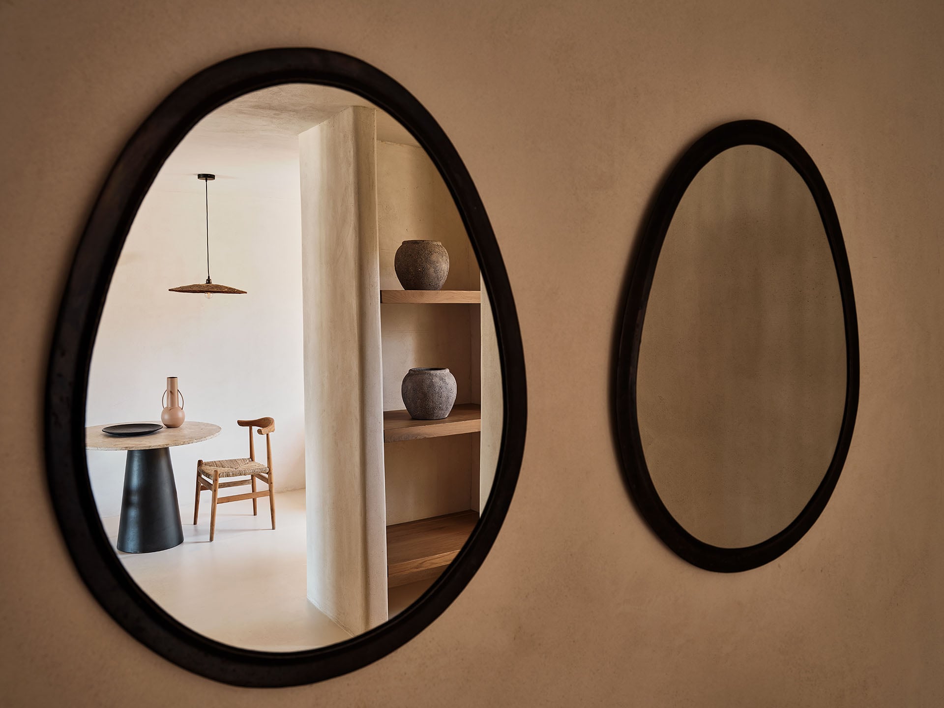 The Wild, Europe — Two black-framed mirrors on warm taupe wall reflecting minimalist dining space with wooden furniture, pendant light, and ceramic vessels on shelving.