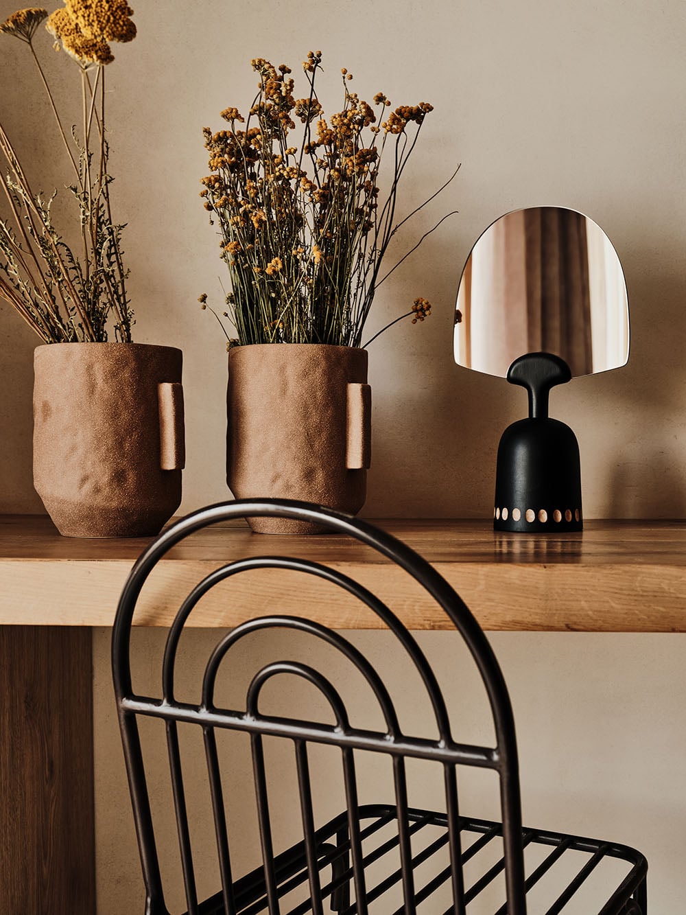 The Wild, Europe — Minimalist hospitality interior with dried florals in cork vessels, black metal chair, and round mirror on warm neutral wall.