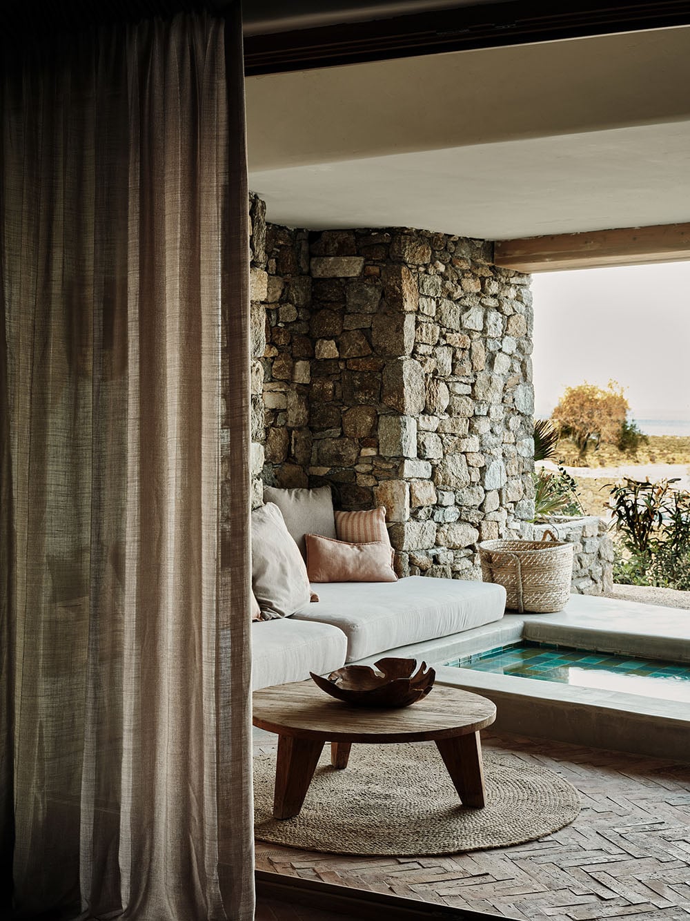 Stone-walled poolside lounge at The Wild with curved seating, wooden table, and linen curtains framing Mediterranean landscape.