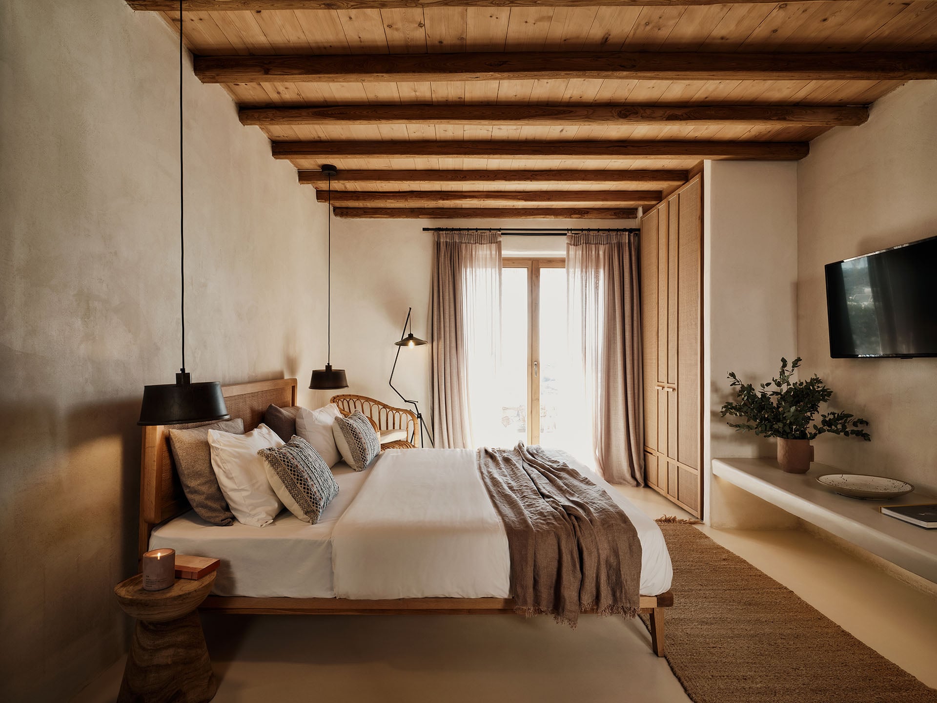 The Wild, Europe — Minimalist bedroom with exposed wooden ceiling beams, natural linen bedding, pendant lighting, and floor-to-ceiling doors opening to landscape views.