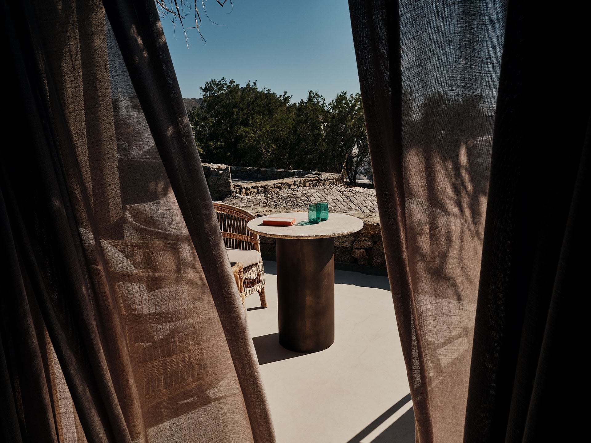 The Wild, Europe — Shaded terrace with linen curtains, cylindrical stone table, woven chair, and Mediterranean landscape beyond stone walls.