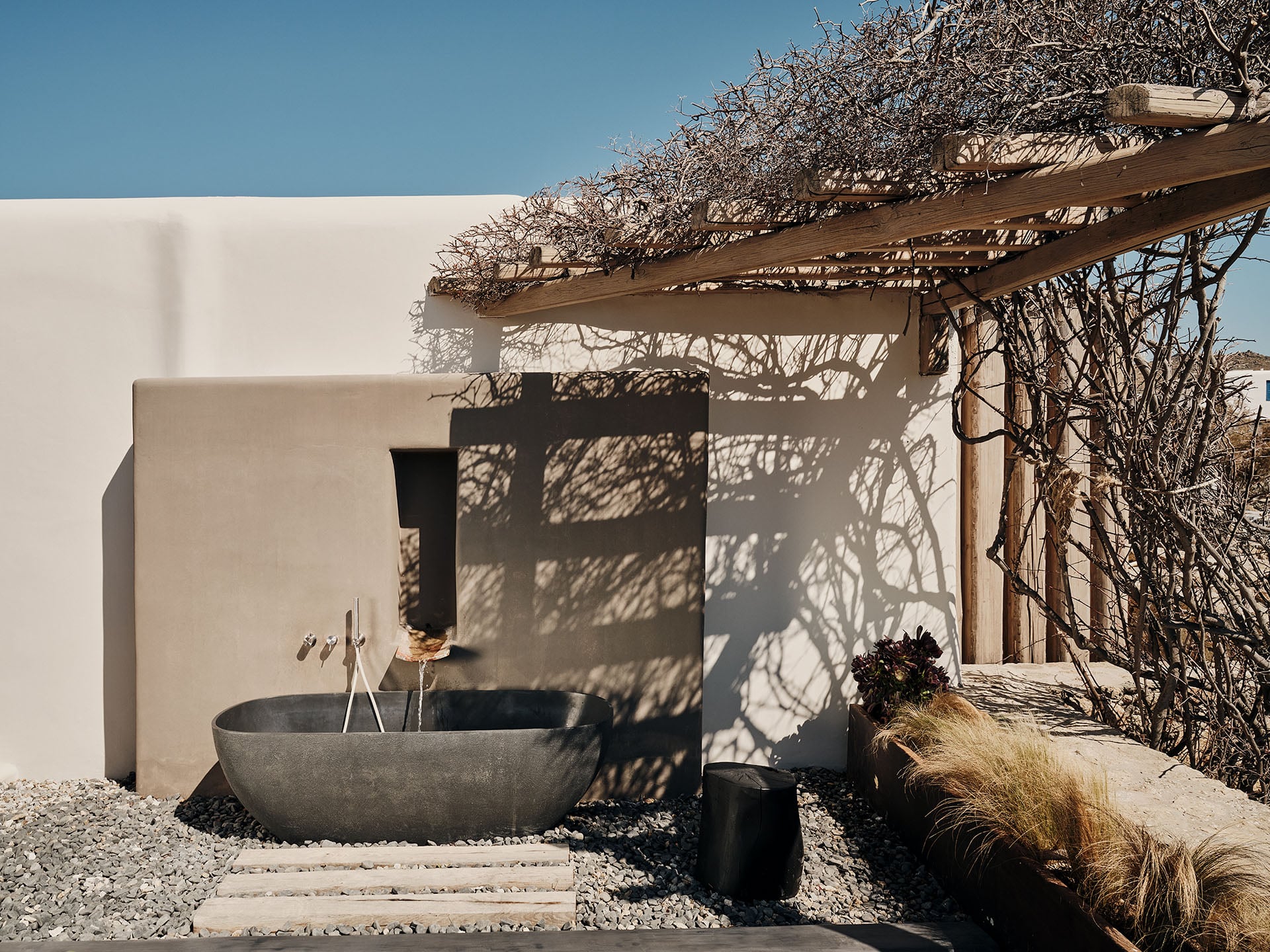 The Wild, Europe — Minimalist outdoor bathroom with concrete soaking tub, wooden pergola, and dry vegetation under clear sky, architectural hospitality design.