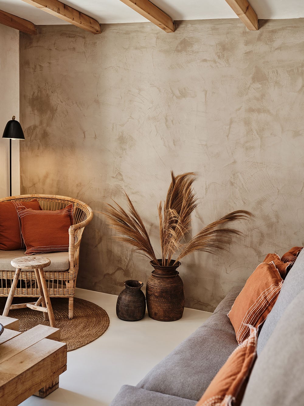 The Wild, Europe — Minimalist hotel suite with textured plaster walls, rattan chair, terracotta accents, dried pampas grass, wooden beams, and warm ambient lighting.