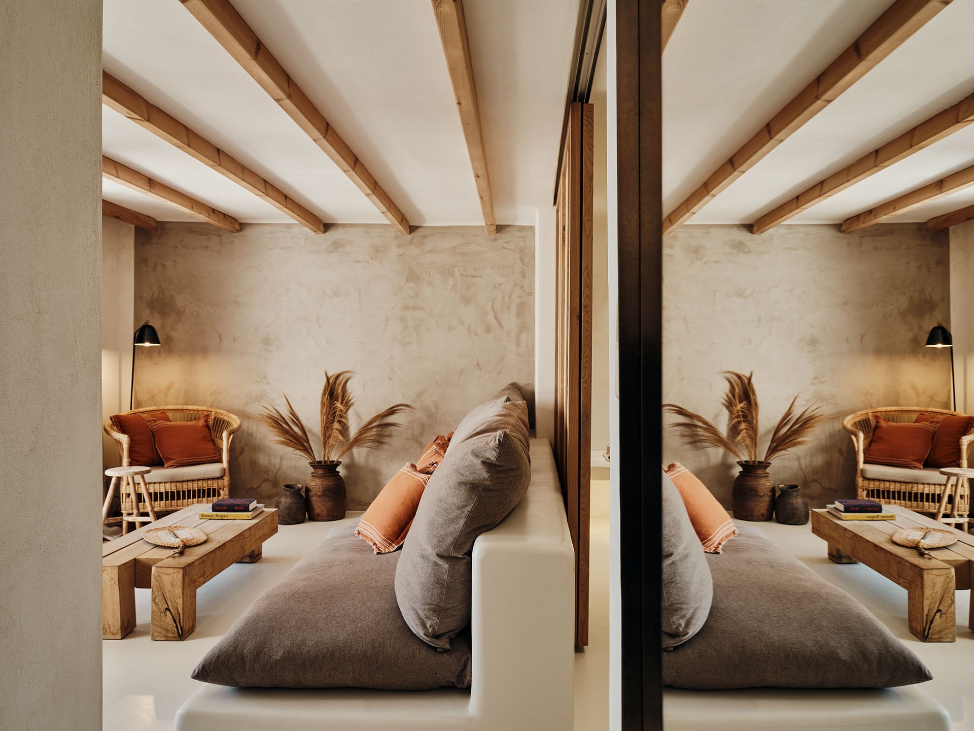 The Wild, Europe — Mirrored spa room with exposed wooden beams, neutral plaster walls, gray floor cushion, woven chair, dried pampas grass, and warm pendant lighting.