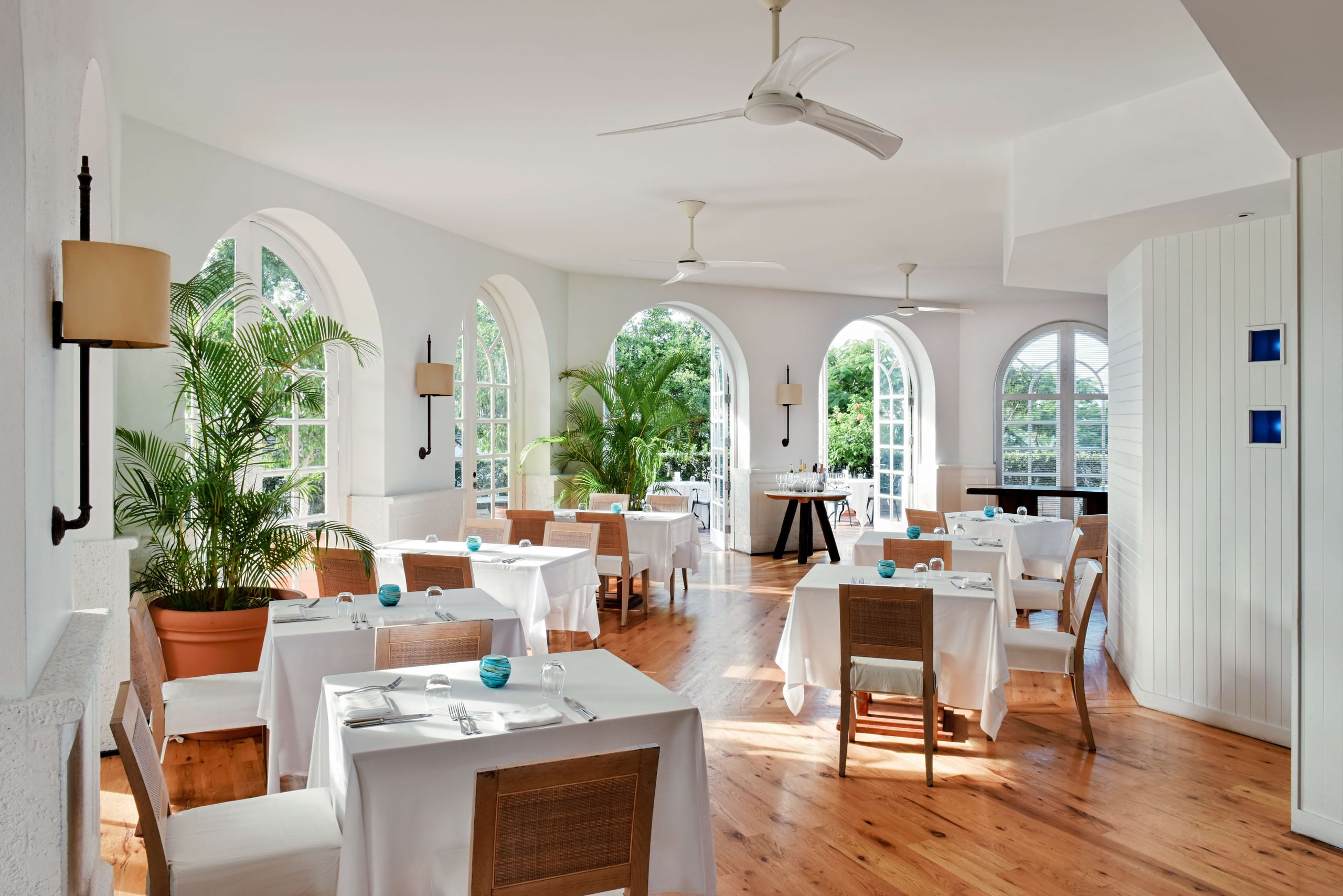 COMO Parrot Cay, North America — Bright dining room with arched windows, woven rattan chairs, white tablecloths, potted palms, wooden floors, and natural light.