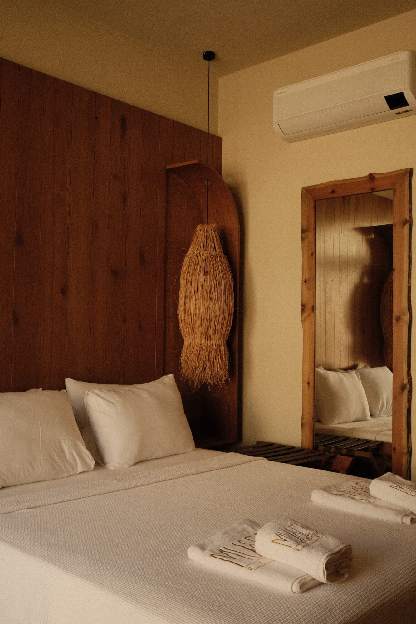 Villa Thalia by Muses Kalkan, Asia — Minimalist bedroom with dark wood headboard, woven pendant light, cream bedding, wooden-framed mirror, air conditioning unit, warm neutral palette.