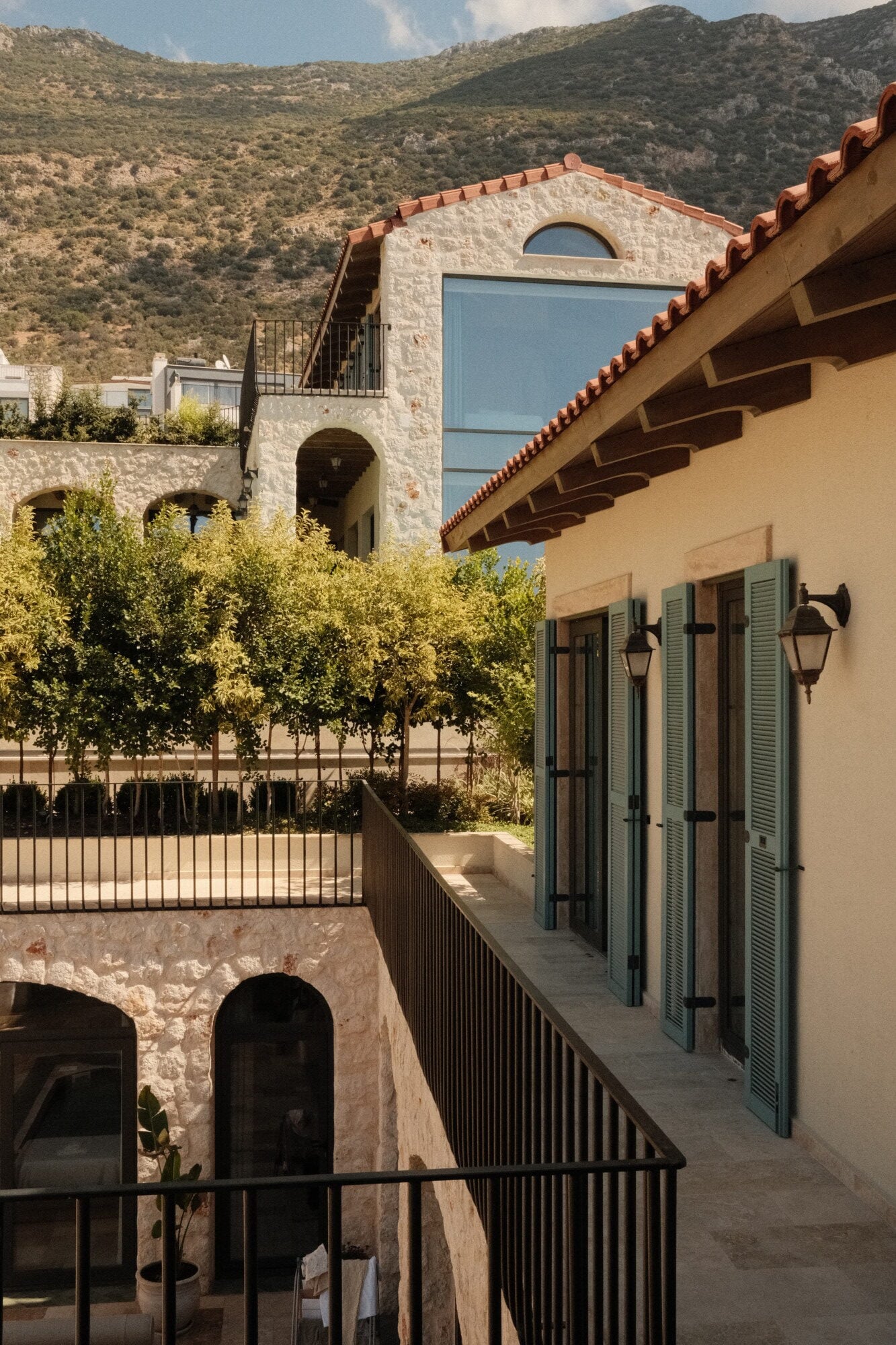 Villa Thalia by Muses Kalkan, Kalkan, Turkey — Terraced courtyard with stone archways, blue shutters, olive tree, and mountain vista under Mediterranean sun.