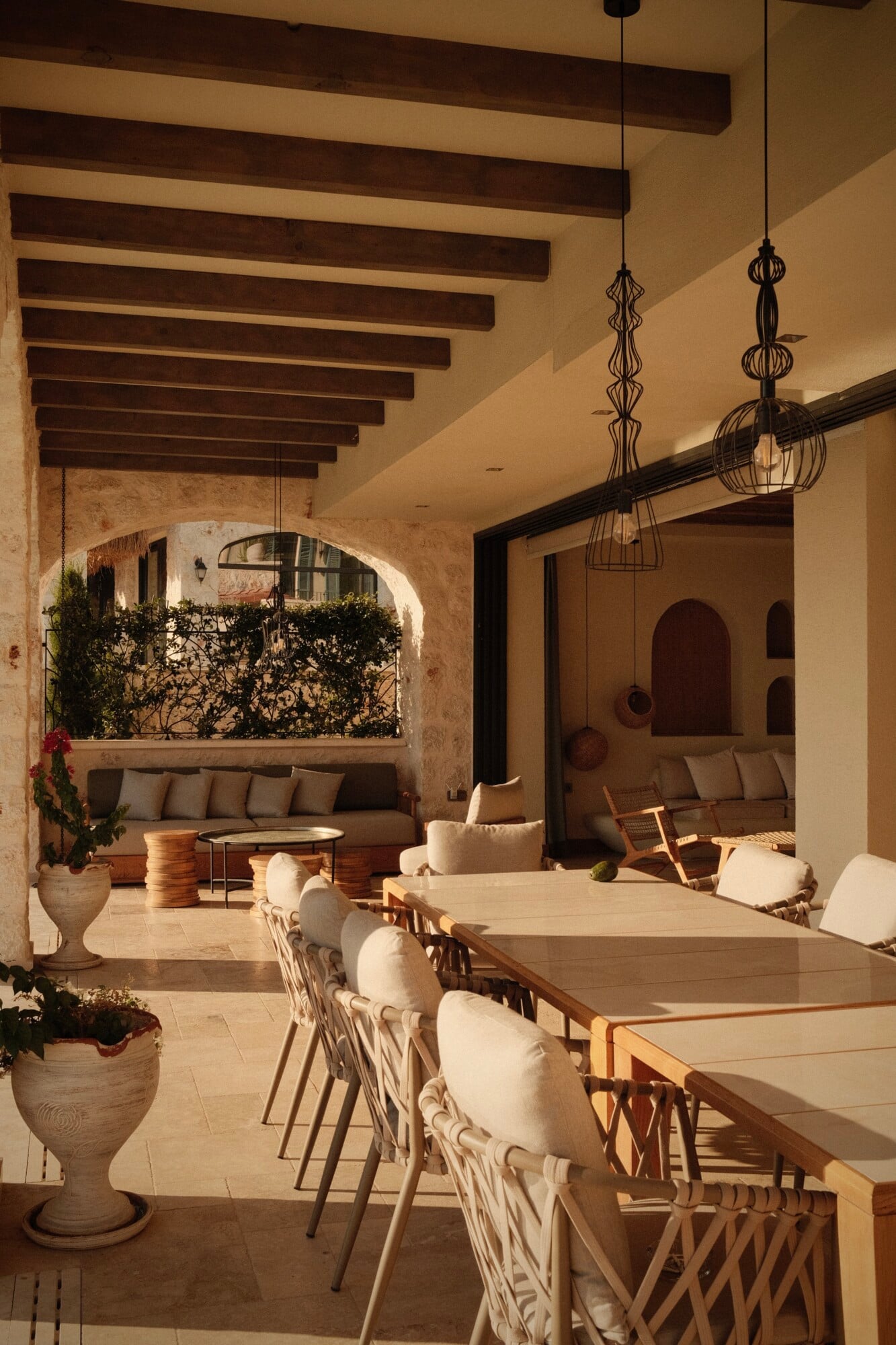Villa Thalia by Muses Kalkan, Asia — Terracotta-tiled courtyard with exposed stone archway, dark wooden beams, cream dining table, woven pendant lights, and seamless indoor-outdoor living space overlooking lounge area.