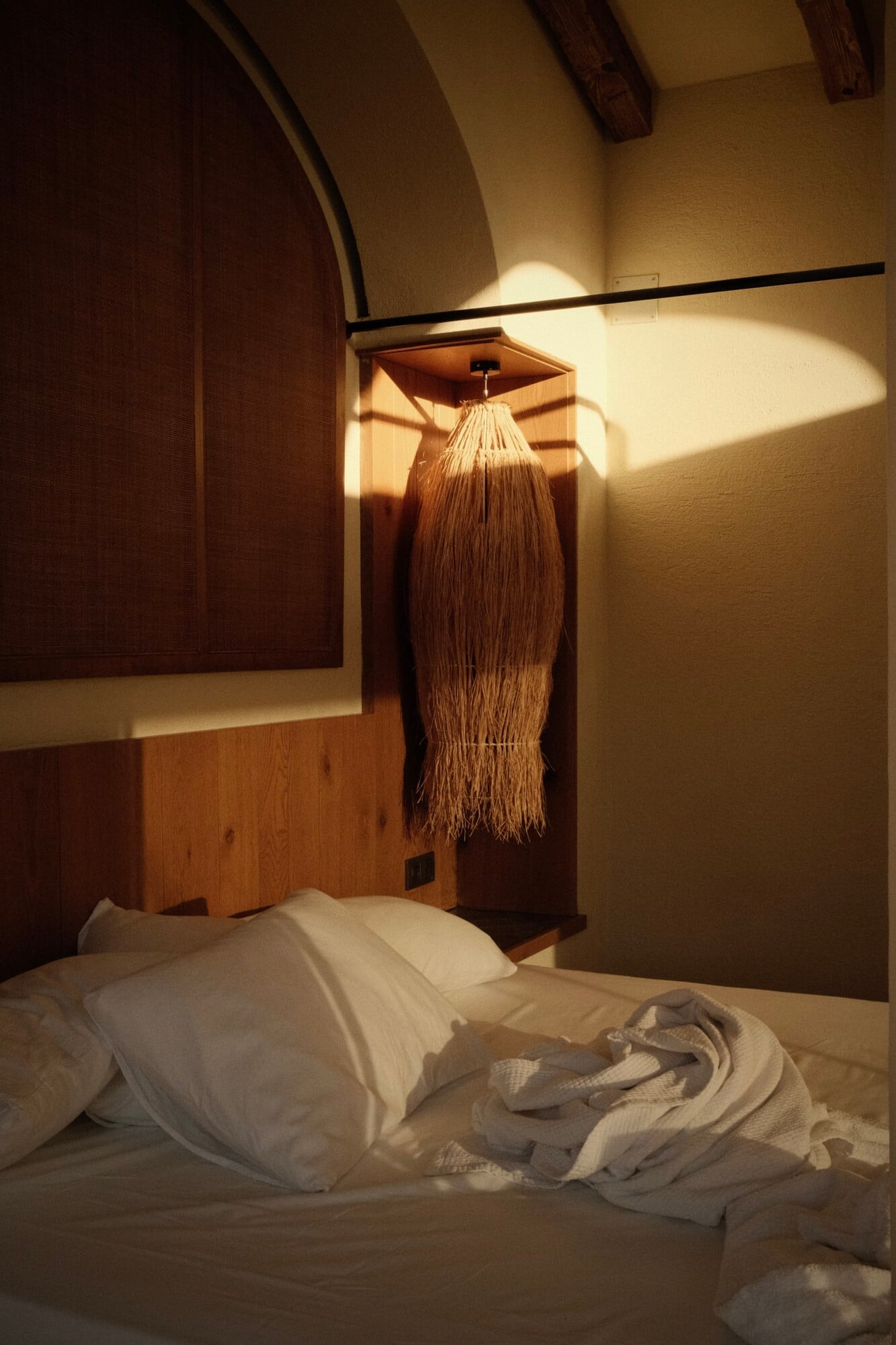 Villa Thalia by Muses Kalkan, Asia — Minimalist bedroom with woven pendant lamp casting warm light over white linens, wooden headboard, and vaulted ceiling with exposed beams.