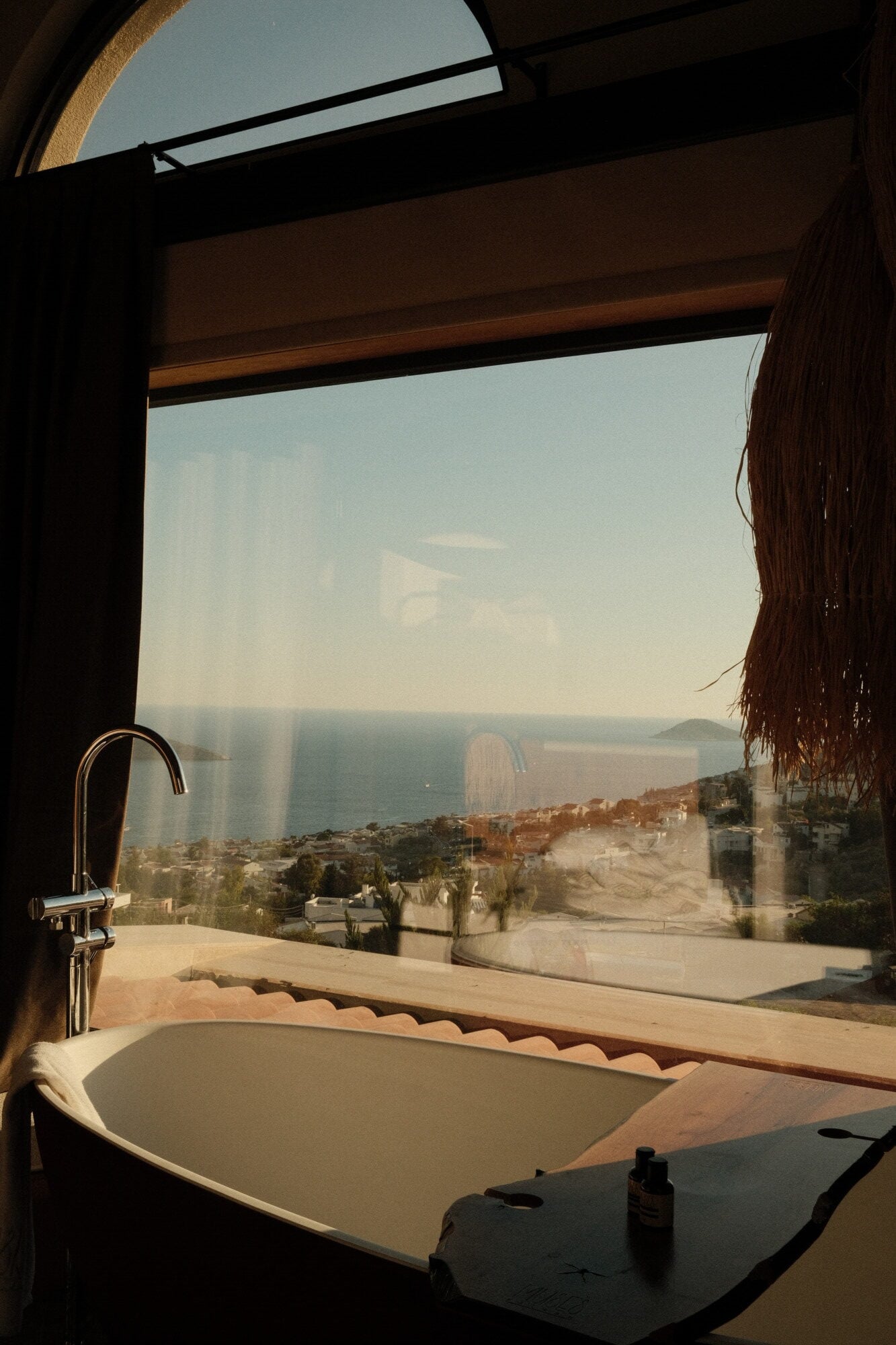 Villa Thalia by Muses Kalkan, Kalkan, Turkey — Modern freestanding bathtub with chrome faucet overlooking Mediterranean coastline through floor-to-ceiling windows, minimalist bathroom design with terracotta tile accents and coastal views.