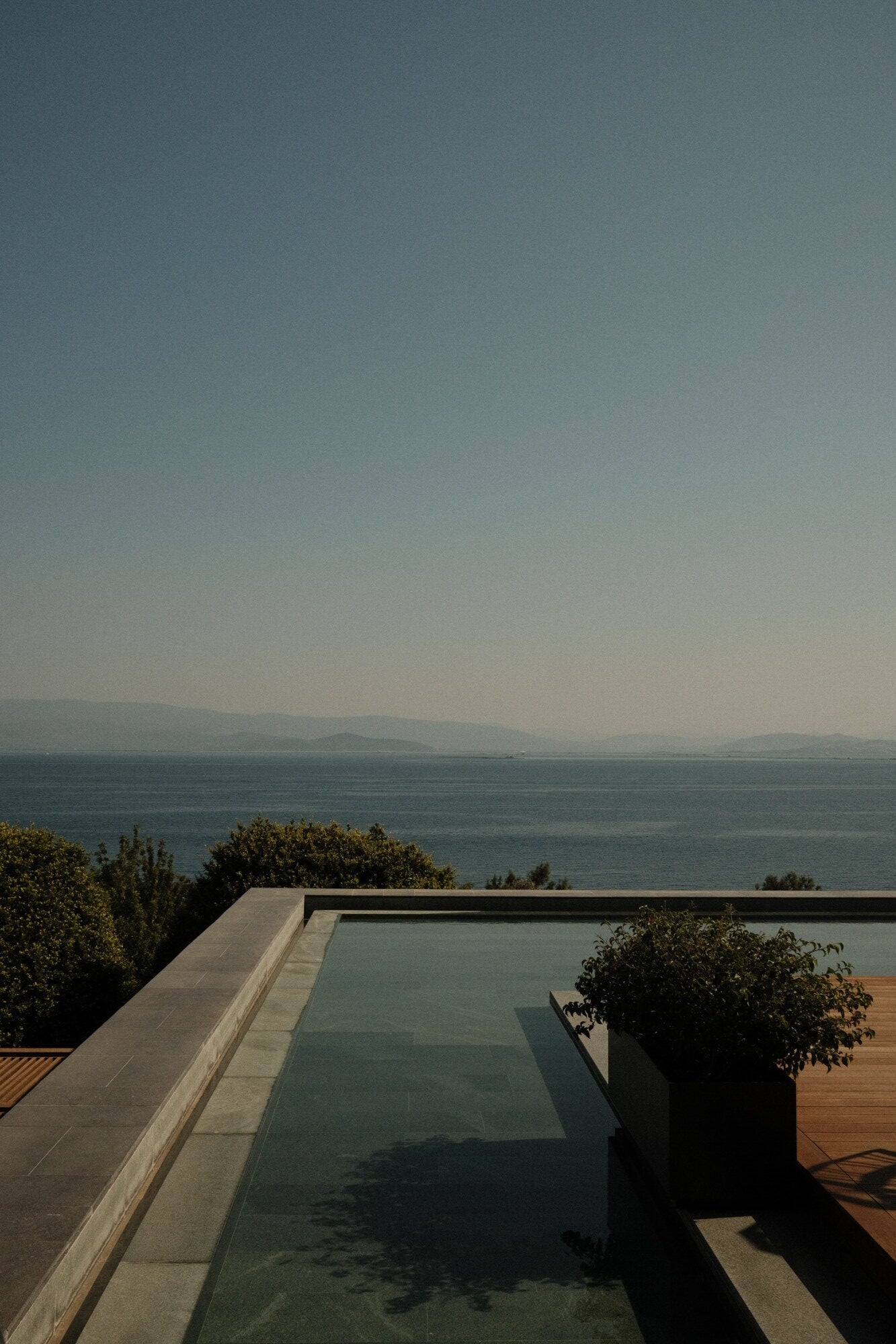 Mandarin Oriental Bodrum, Asia — Infinity pool with glass edge overlooking Aegean Sea, minimalist stone terrace, hazy coastal mountains at horizon.