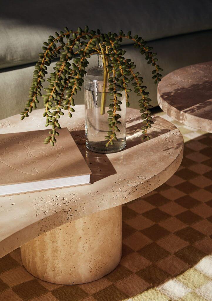 Kisawa Sanctuary, Africa — Travertine round table with cascading string-of-pearls succulent in glass vase beside leather-bound book, dappled sunlight across patterned floor.