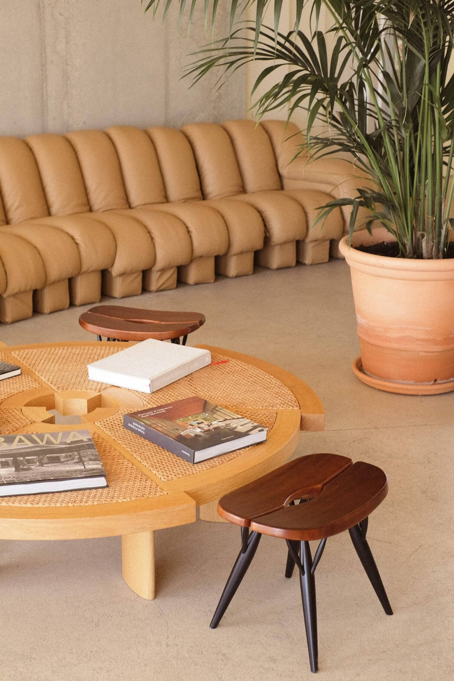 Les Roches Rouges, Europe — Curved caramel leather sofa, wooden coffee table with woven top, dark wood stools, potted palm, minimalist hospitality interior design.