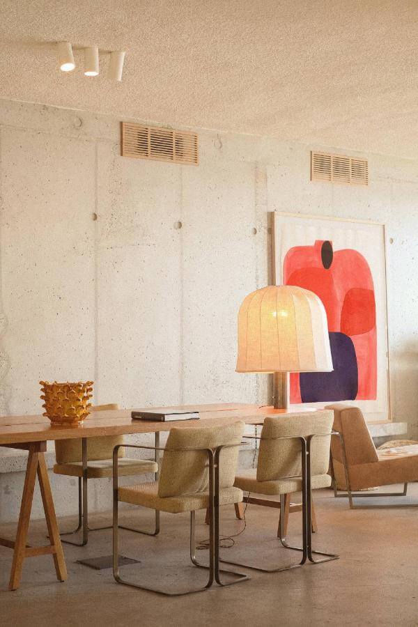 Les Roches Rouges, Europe — Minimalist dining room with wooden table, tan chairs, pendant lamp, concrete walls, and abstract red artwork.