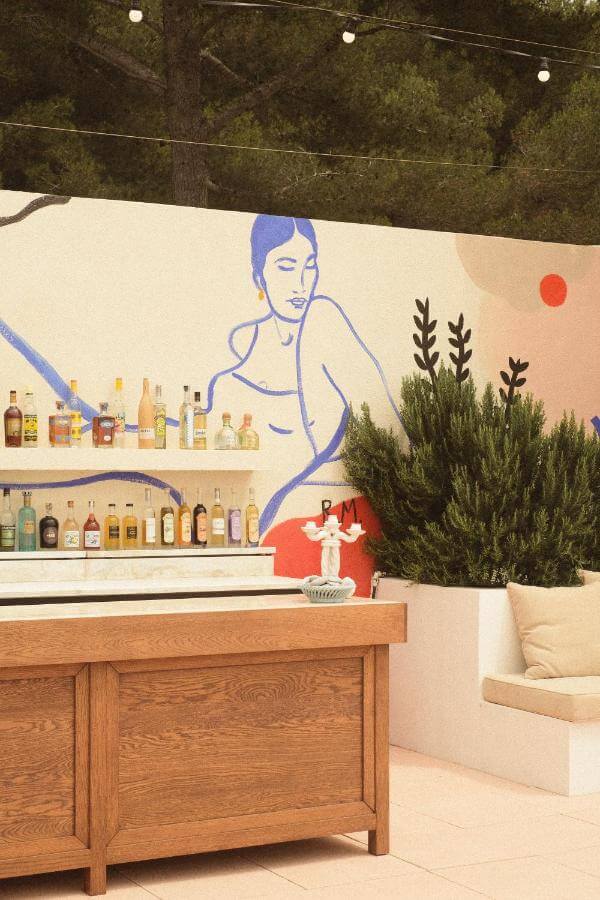 Les Roches Rouges, Europe — Outdoor bar with blue-line figure mural, wooden counter, bottle display, potted cypress plants, and earth-tone architectural backdrop.