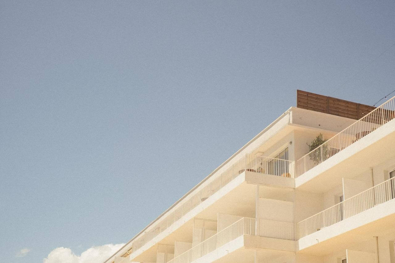 Les Roches Rouges, Europe — Minimalist cream-colored hotel facade with metal railings and wooden privacy screen, clear blue sky, contemporary hospitality architecture.