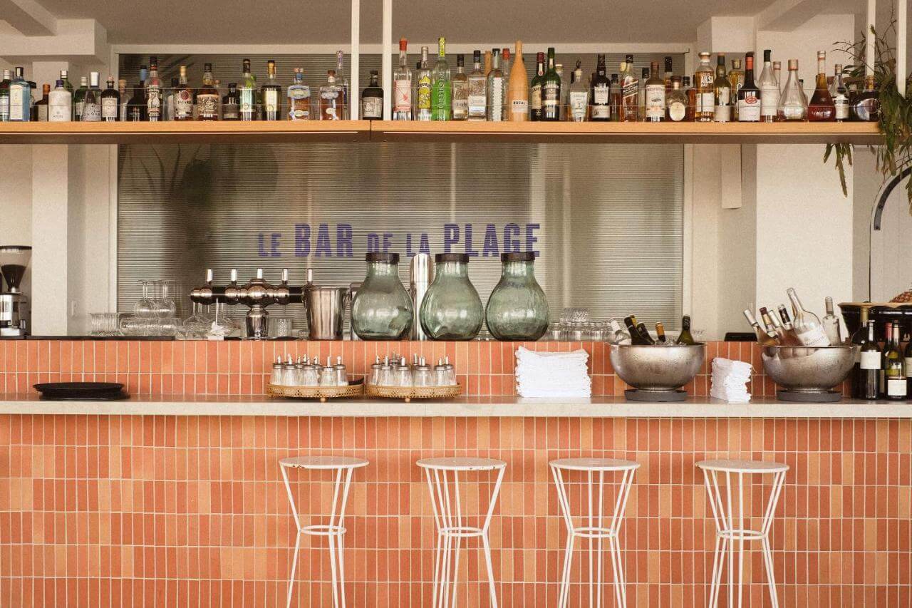 Le Bar de La Plage, Europe — Terracotta tile bar counter with white metal stools, brass vessels, glass bottles on wooden shelving, minimalist coastal hospitality design.