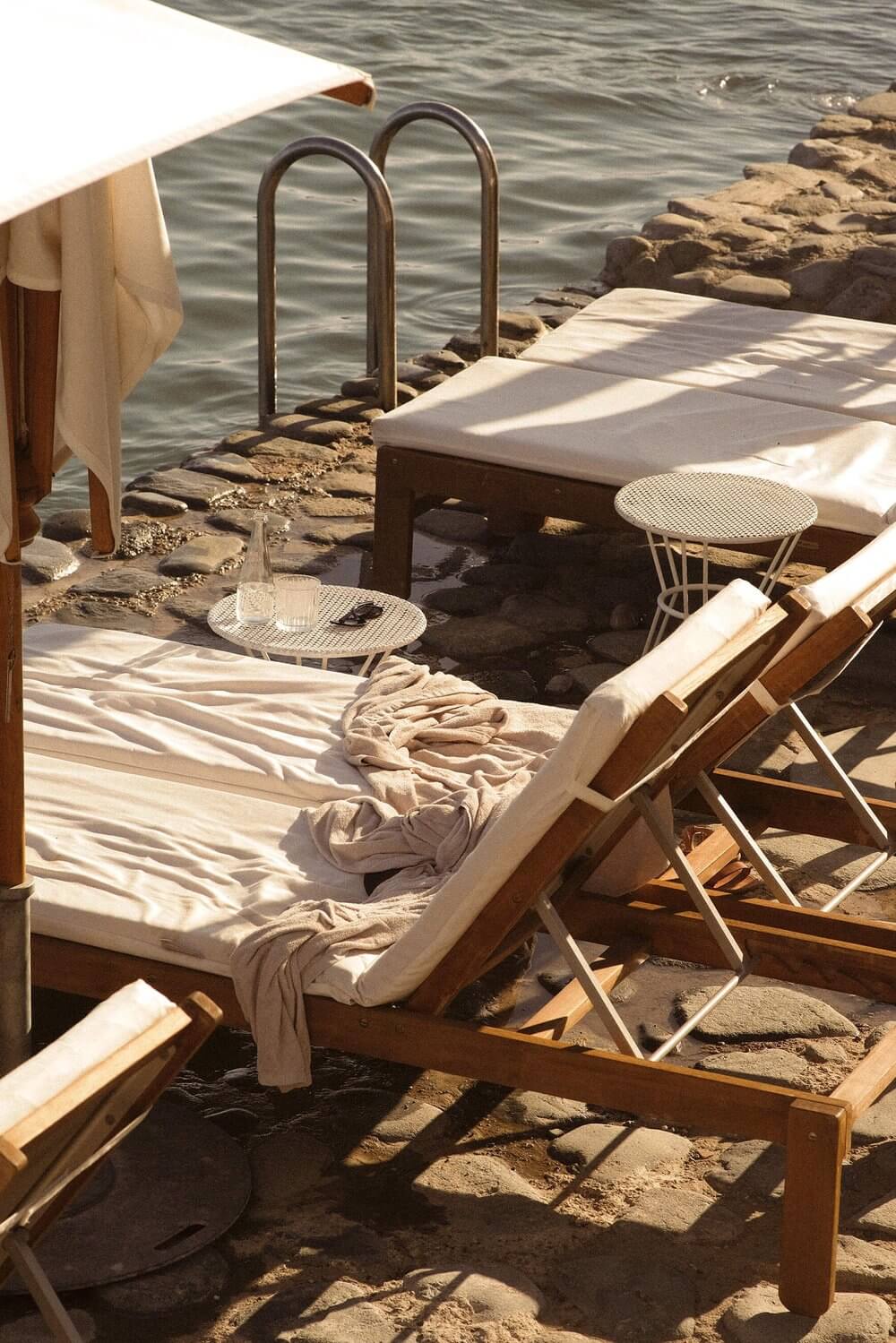 Les Roches Rouges, Europe — Sunlit coastal terrace with canvas canopy, wooden loungers, metal side tables, and stepped stone access to calm water.