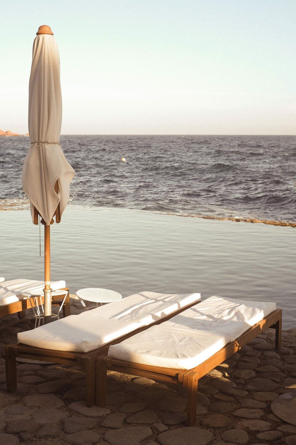 Les Roches Rouges, Europe — Wooden loungers with white cushions beneath closed beige umbrella on sandy beach at sunset, Mediterranean sea beyond.