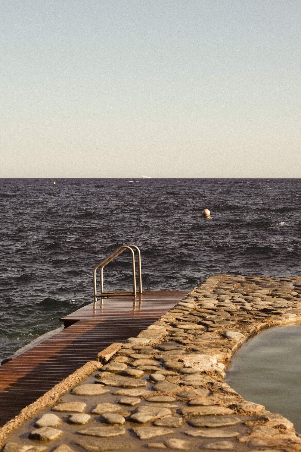Les Roches Rouges, Europe — Wooden dock with metal ladder extending into dark sea, stone breakwater, minimalist coastal architecture at dusk.