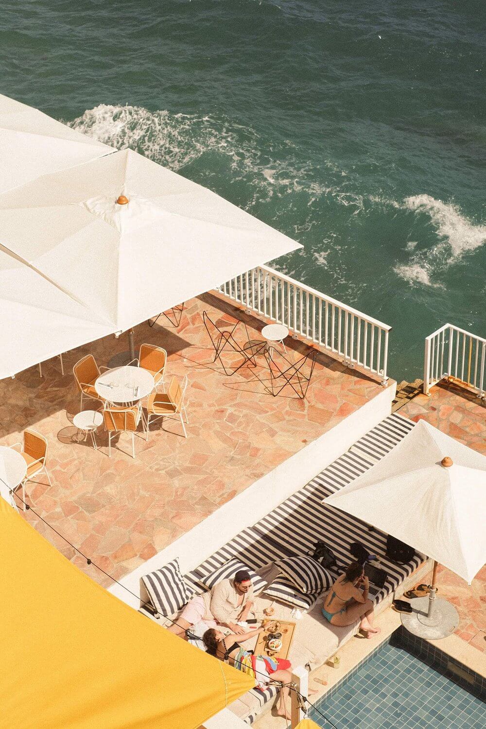 Les Roches Rouges, Europe — Aerial view of clifftop terrace with striped loungers, white umbrellas, and guests overlooking turquoise Mediterranean waves below.