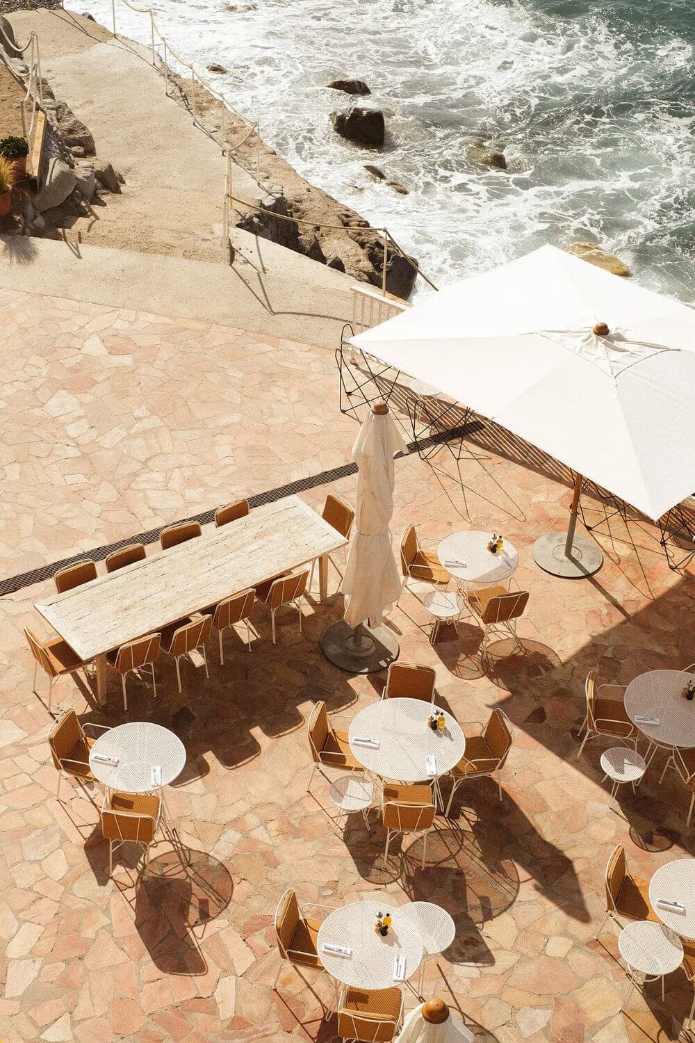 La Plage, Europe — Terracotta coastal terrace with wooden chairs, white umbrellas, and dining tables overlooking Mediterranean waves and rocky shoreline.