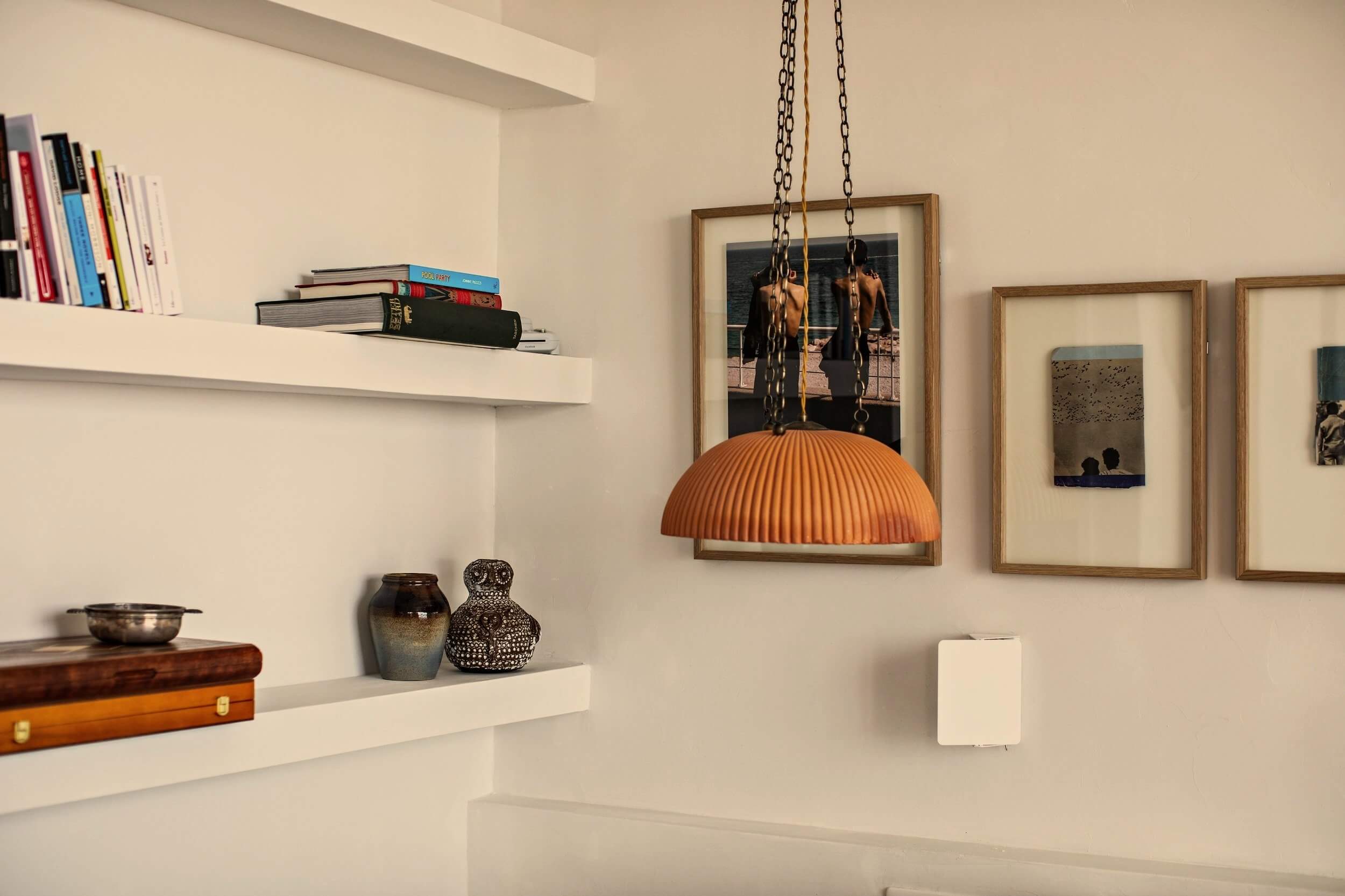 Les Roches Rouges, Europe — Minimalist gallery wall with floating shelves, stacked books, framed photographs, pendant lamp with pleated orange shade, and decorative objects in neutral interior.