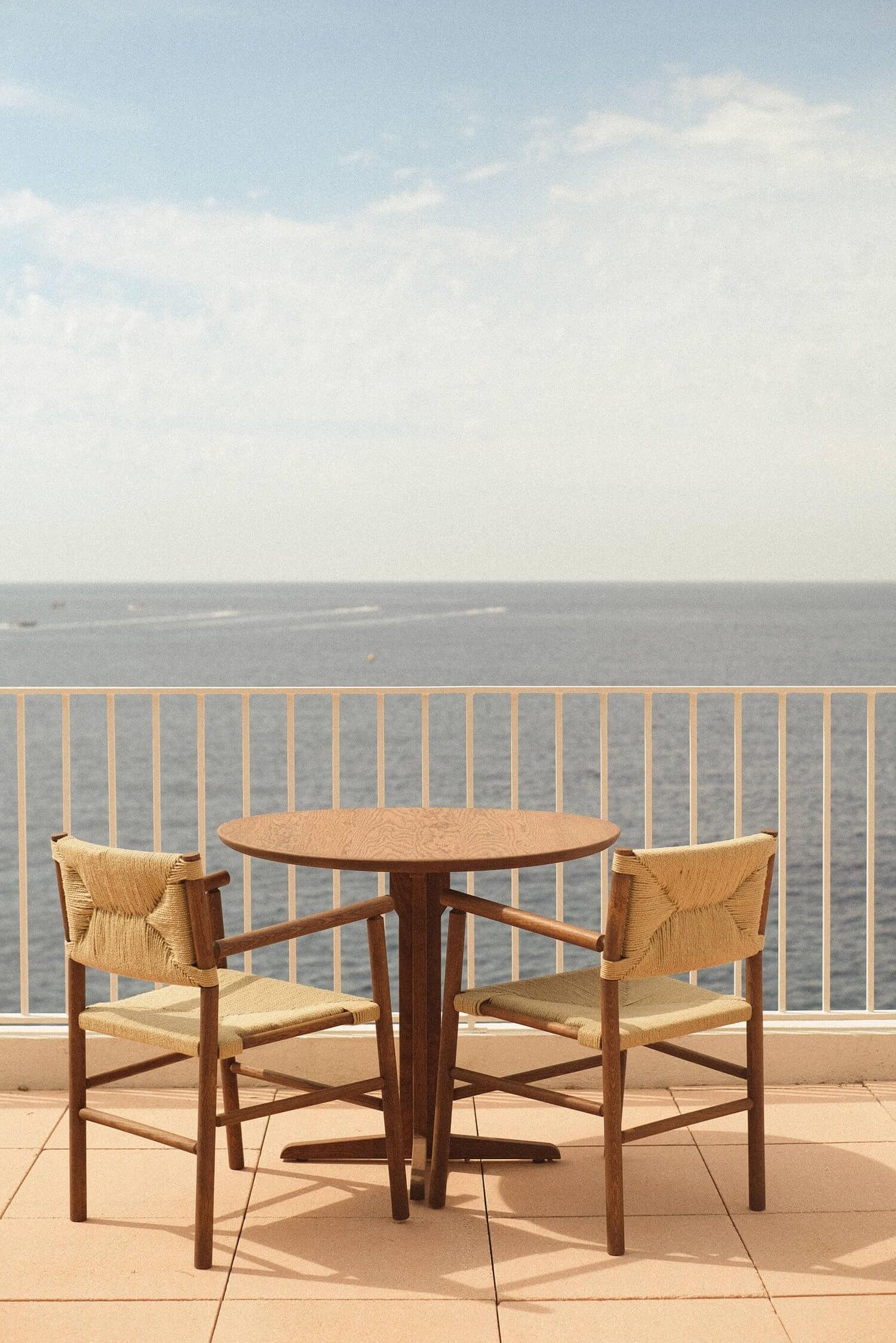 Les Roches Rouges, Europe — Wooden round table and woven chairs on coastal terrace overlooking calm sea, morning light, minimalist seaside dining.
