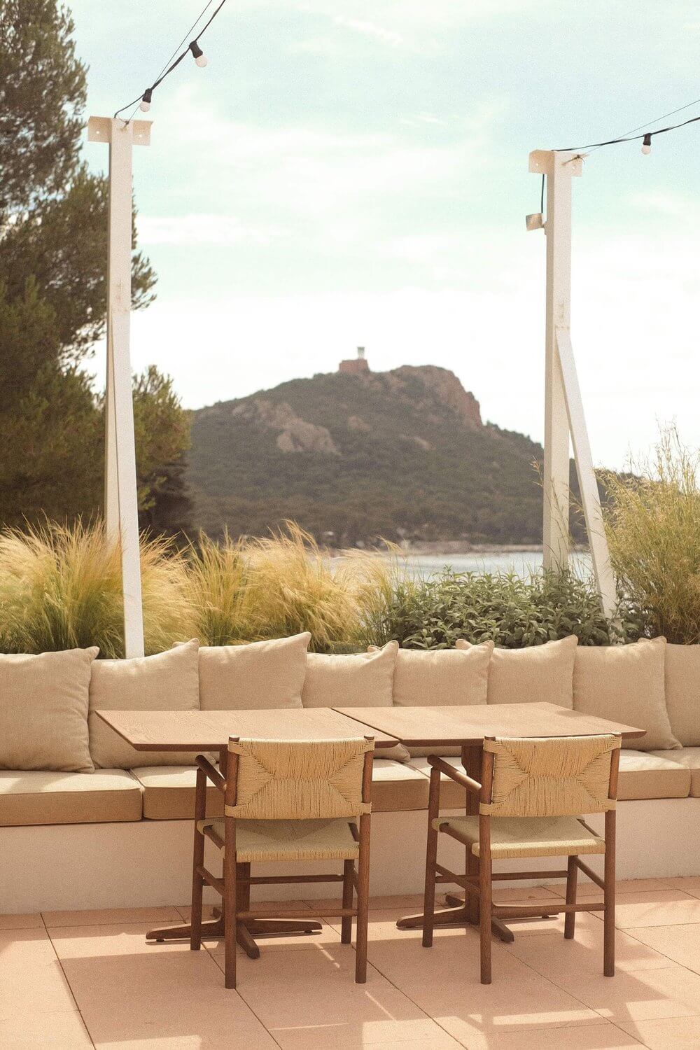 Récif, Europe — Waterfront lounge with cream built-in seating, woven wooden chairs, pergola, ornamental grasses, and mountain lake views at dusk.