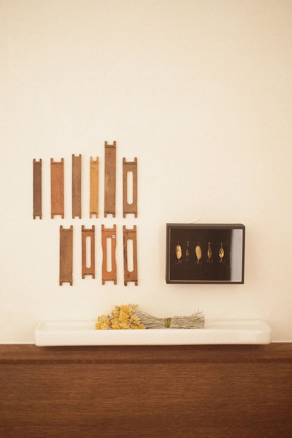 Les Roches Rouges, Europe — Vintage fishing lures and wooden loom parts displayed on cream wall above white tray with dried flowers on wood shelf.