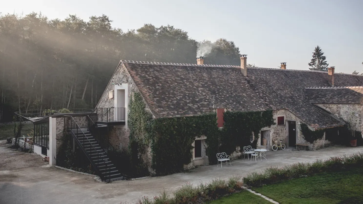 Le Barn, Europe — Stone barn complex with slate roofs and ivy-covered walls at dawn, featuring modern metal staircase and courtyard seating amid misty woodland.