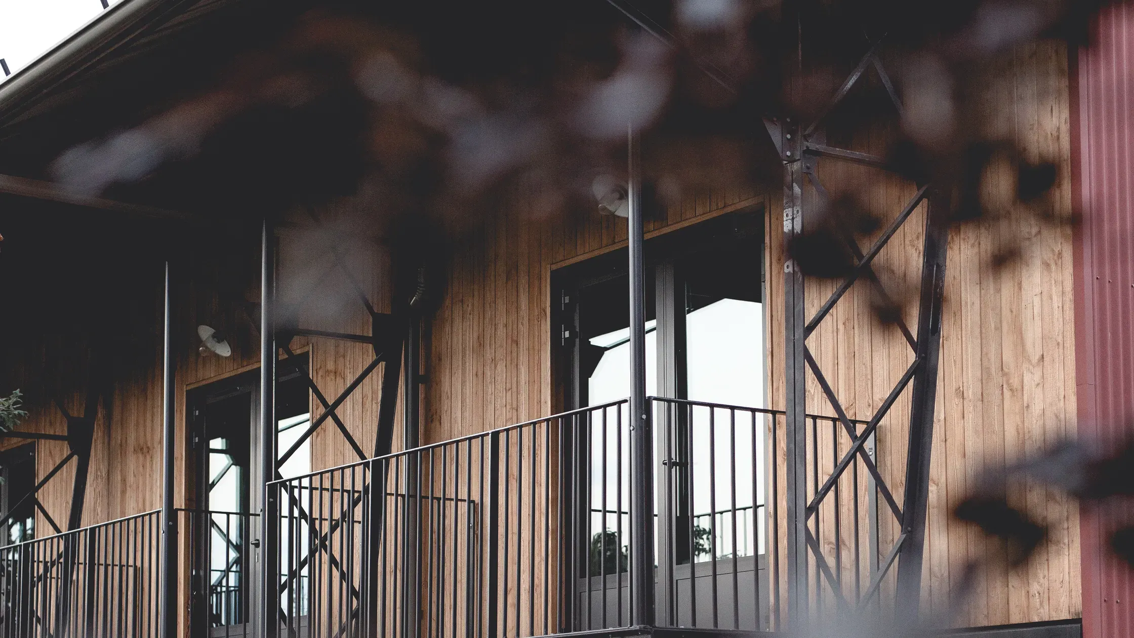 Le Barn, Europe — Modern barn interior with weathered wood siding, black metal railings, floor-to-ceiling glass doors opening onto countryside vista, dappled shadow patterns.