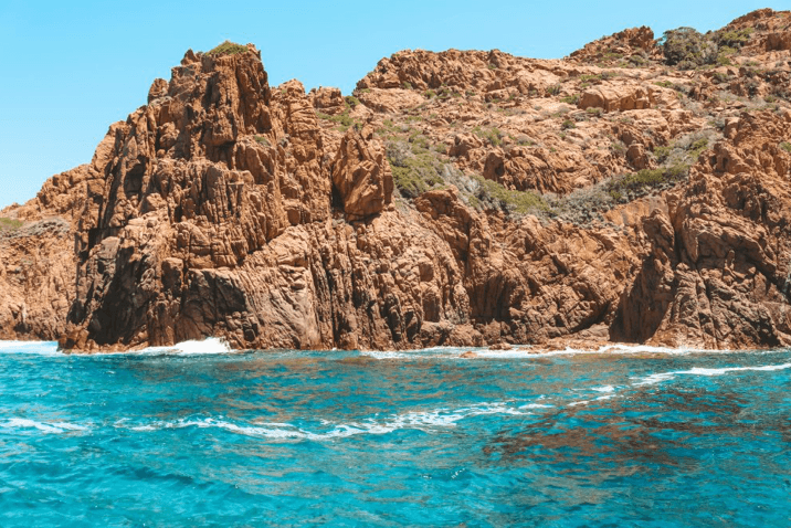 Les Calanques de Piana, Corsica, France — Dramatic red granite cliffs plunging into turquoise Mediterranean waters with white waves and sparse vegetation.