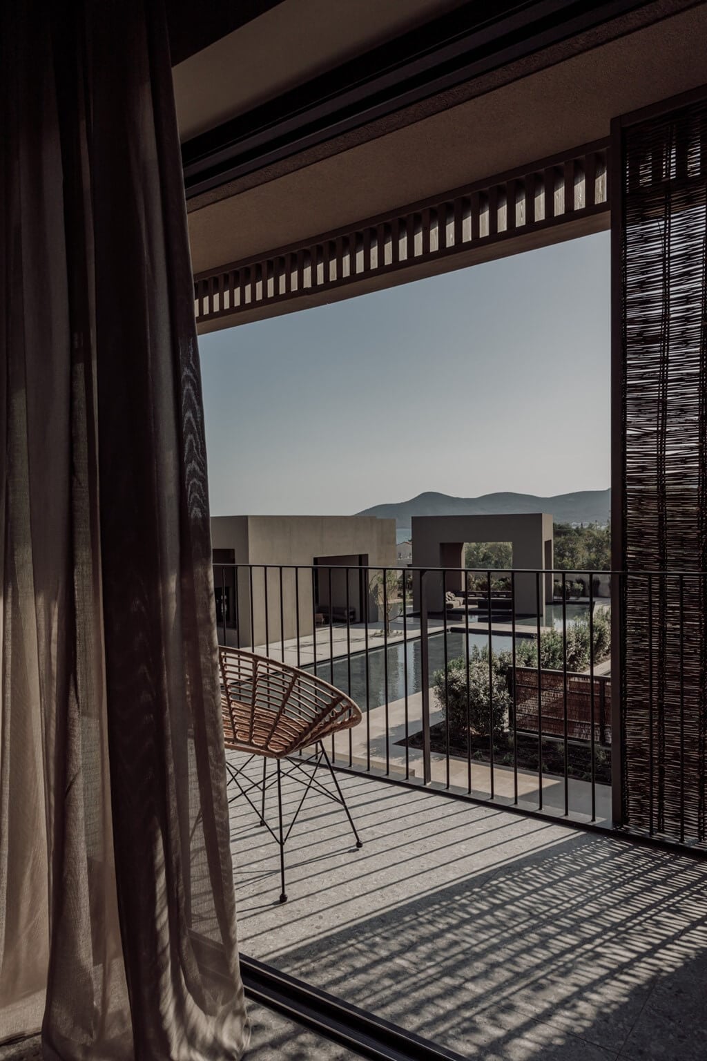 Casa Cook Samos, Greece — Minimalist terrace with woven chair, wooden shutters, mountain views, and afternoon shadow patterns on polished concrete.