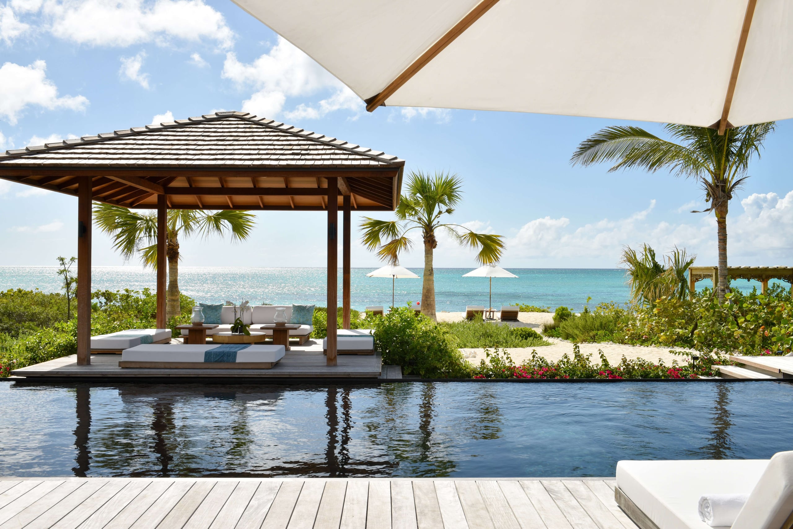COMO Parrot Cay, North America — Waterfront pavilion with teak pergola, lounge seating, infinity pool, and turquoise Caribbean Sea beyond palm-lined beach.