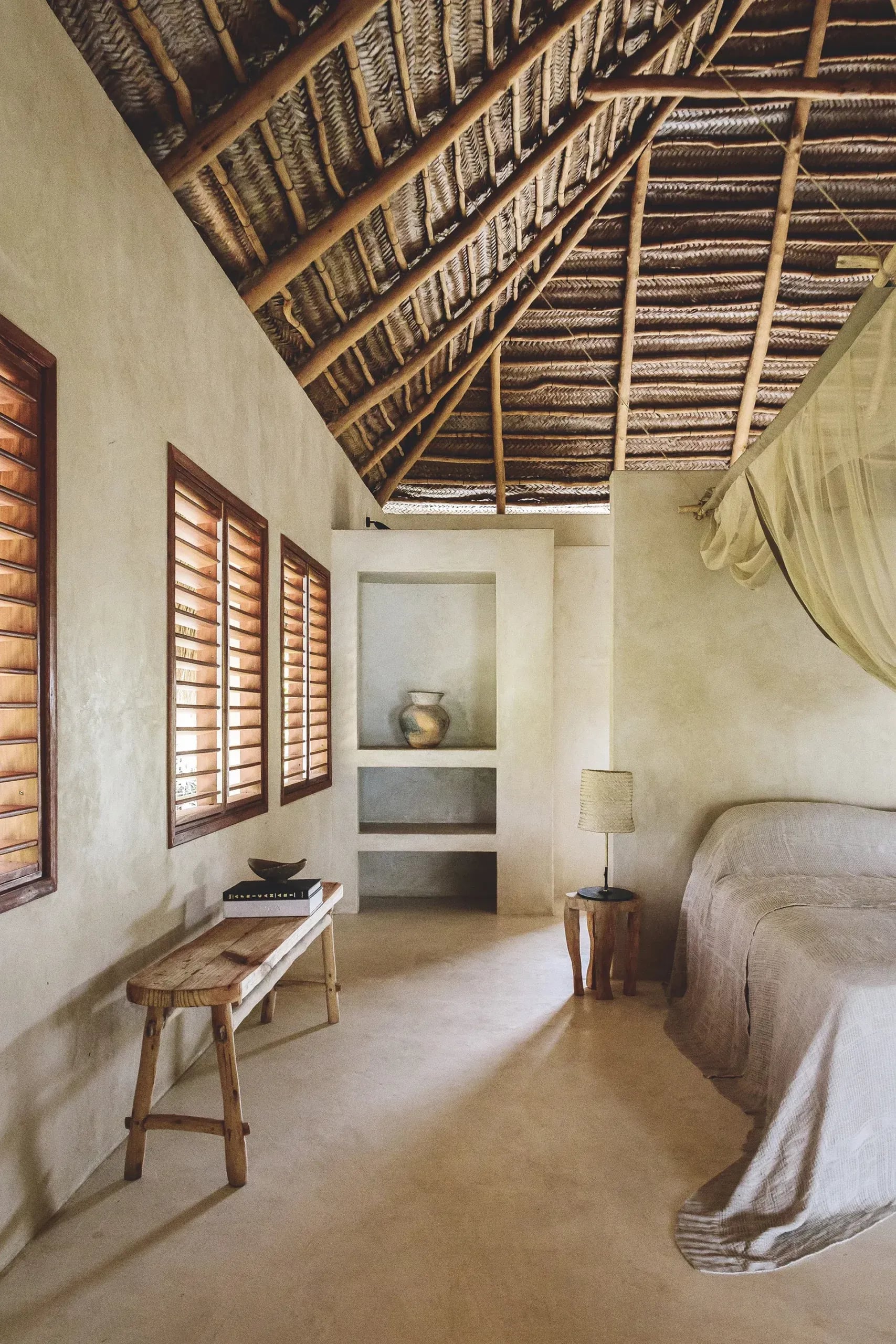Sussurro, Africa — Minimalist bedroom with timber lattice ceiling, wooden shutters, plaster walls, and woven furnishings creating serene colonial aesthetic.
