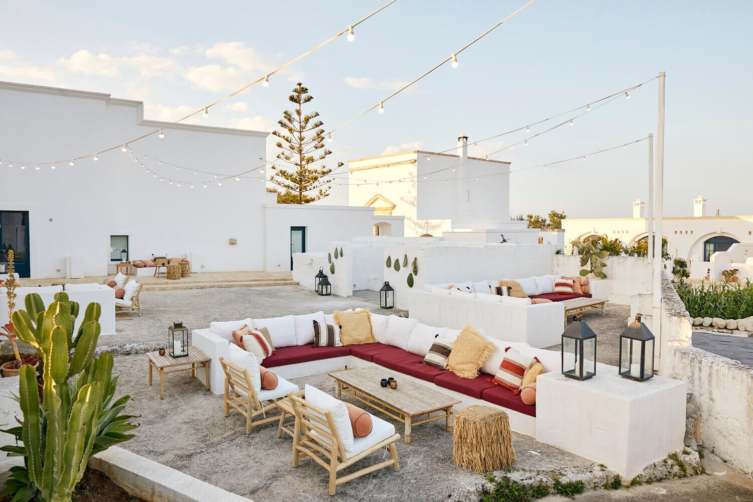 Apero Bar at Masseria Calderisi, Italy — whitewashed courtyard with burgundy sectional, wooden loungers, string lights, cacti, and Moorish architecture at sunset.