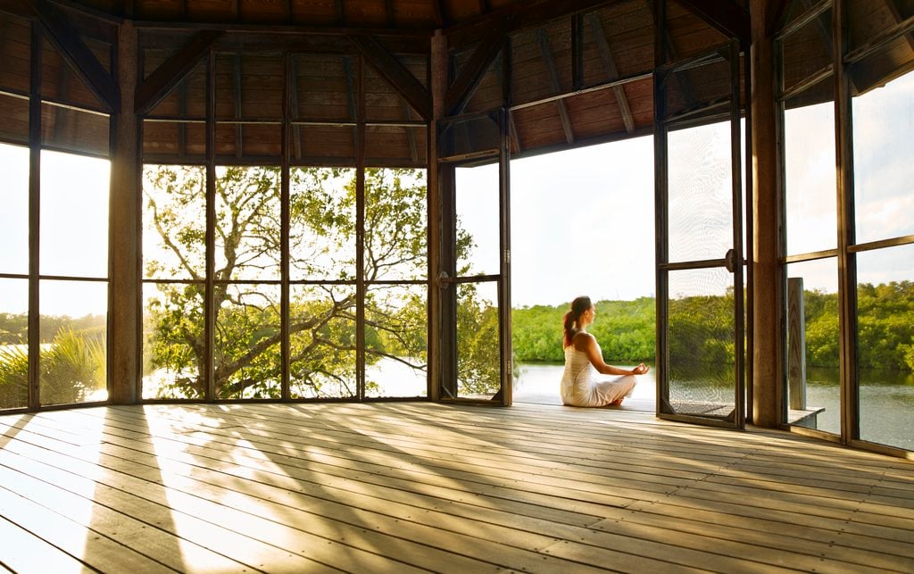 COMO Shambhala, North America — Woman in meditation pose on wooden deck overlooking mangrove river, surrounded by floor-to-ceiling windows and natural light filtering through wooden beams.