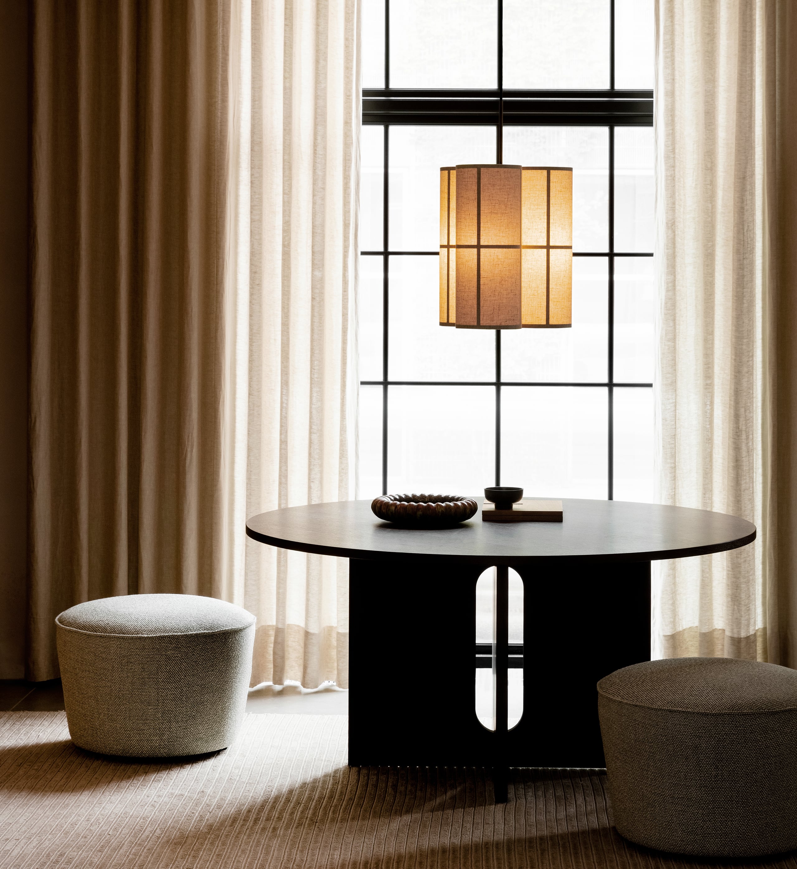 The Audo, Europe — Minimalist dining space with black round table, linen poufs, pendant lamp, and gridded window overlooking garden.