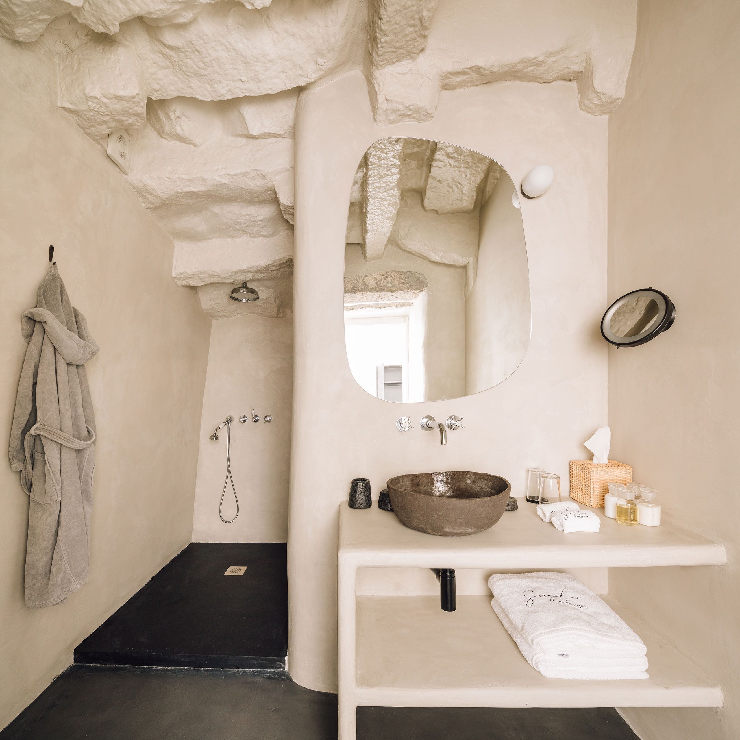 White Hotel, Europe — Minimal bathroom with raw stone walls, curved mirror, earthenware vessel sink, black floor, and neutral linens showcasing contemporary luxury design.